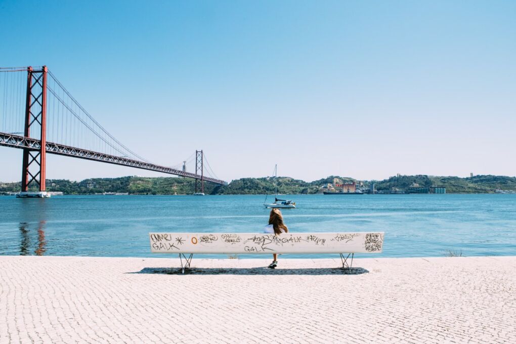 A girl sitting on a bench overlooking the 25 de Abril Bridge and the River Tagus 