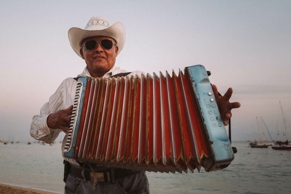 An older Mexican man playing the accordion on the beach. He's wearing sunglasses and a cowboy hat. 