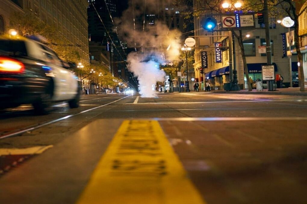 A nighttime city street with a police car driving past on the left and steam coming form a grate in the center of the image.