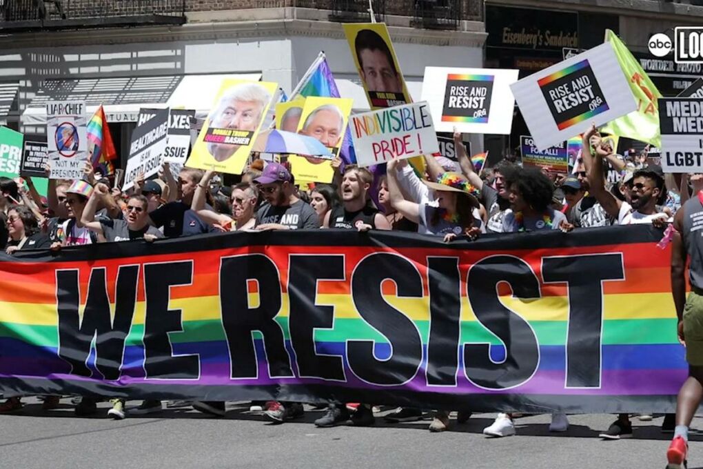 A LGBTQAI+ pride rally holding many protest signs including a large rainbow banner that reads "We resist" in large black letters. 