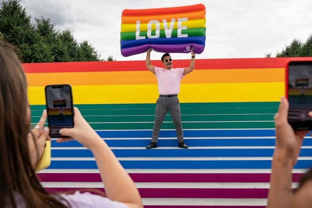 A man standing on rainbow set of steps. He is holding a rainbow inflatable matters with the word "Love" printed on it in big white letters.