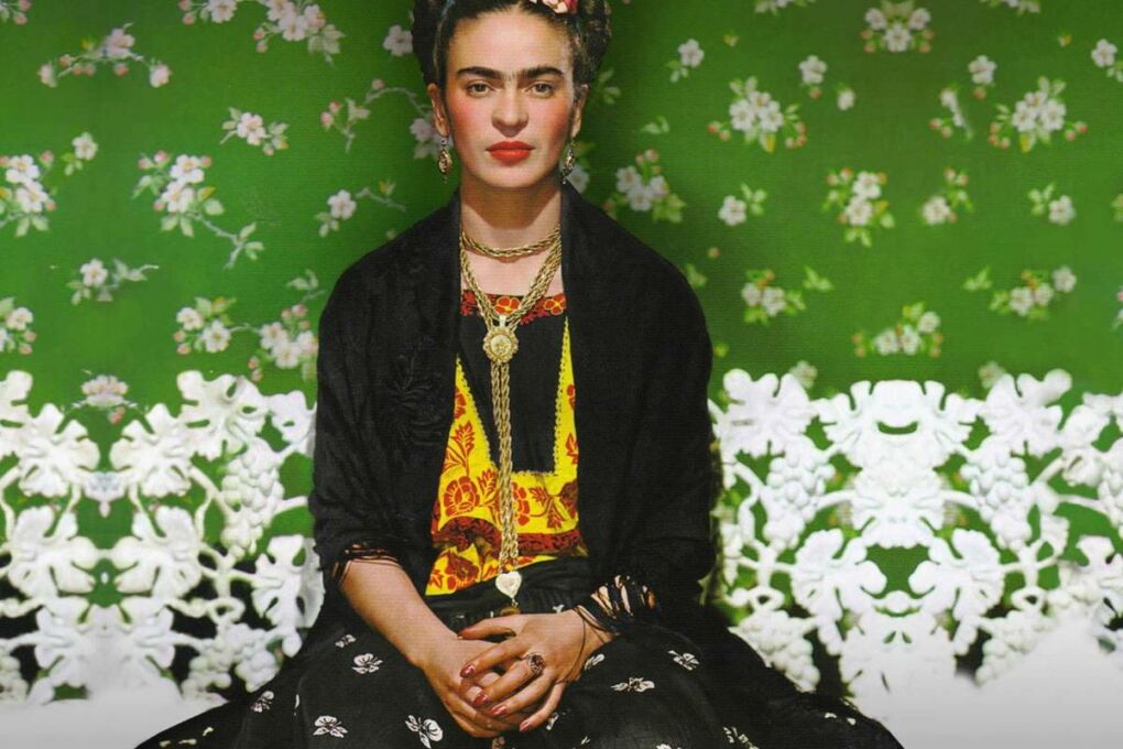 Freda Kahlo sitting on a white chair in front of a green wall with white flowers on it.