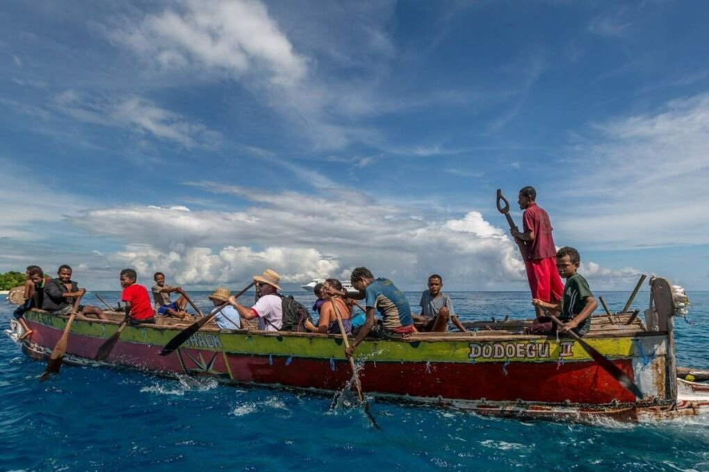 Tourist enjoying a boat tour in Papua New Guinea|A sail boat on the Milne Bay waters|New Guinean tribal people at a festival|A boat sailing across the blue waters of Milne Bay||New Guinean tribal people dancing in their traditional clothes|A view of the Rabaul volcano through the trees|A quiet road through the forests of Papua New Guinea|View of a shoreline in Papua New Guinea|A gorgeous orange sunset over Milne Point waters